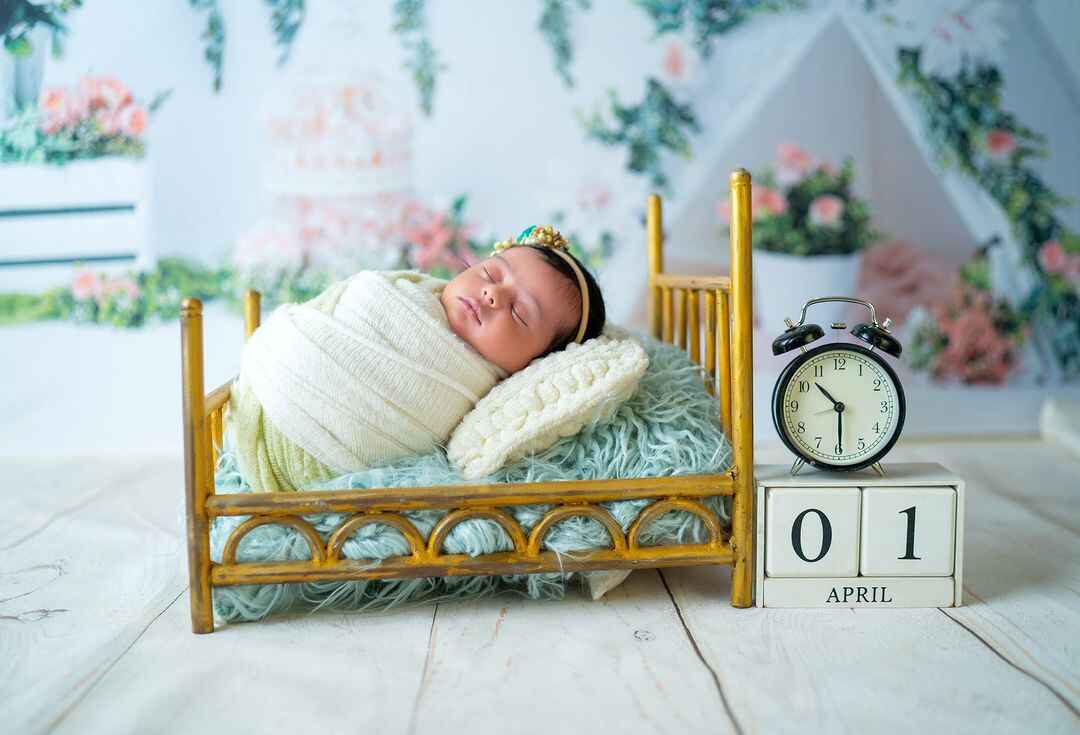 Newborn baby girl in white dress photographed with clock and flower bed setup